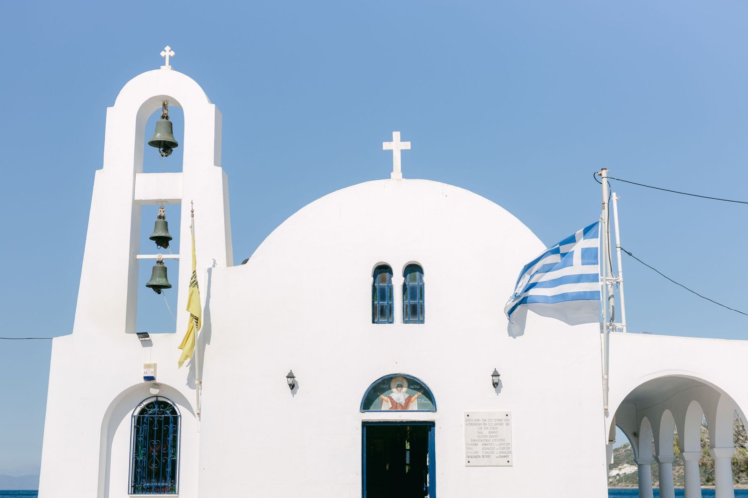 Traditional white Greek Orthodox church with bell tower and blue dome against clear sky, with Greek flag flying in Santorini.