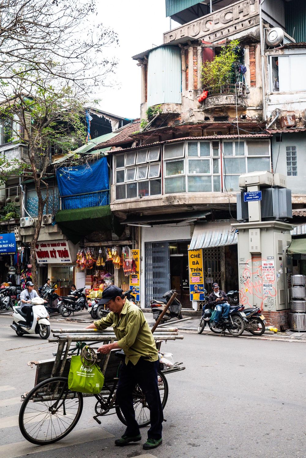Street scene in Hanoi Vietnam showing old apartment building with motorbikes parked below and person with bicycle on sidewalk.