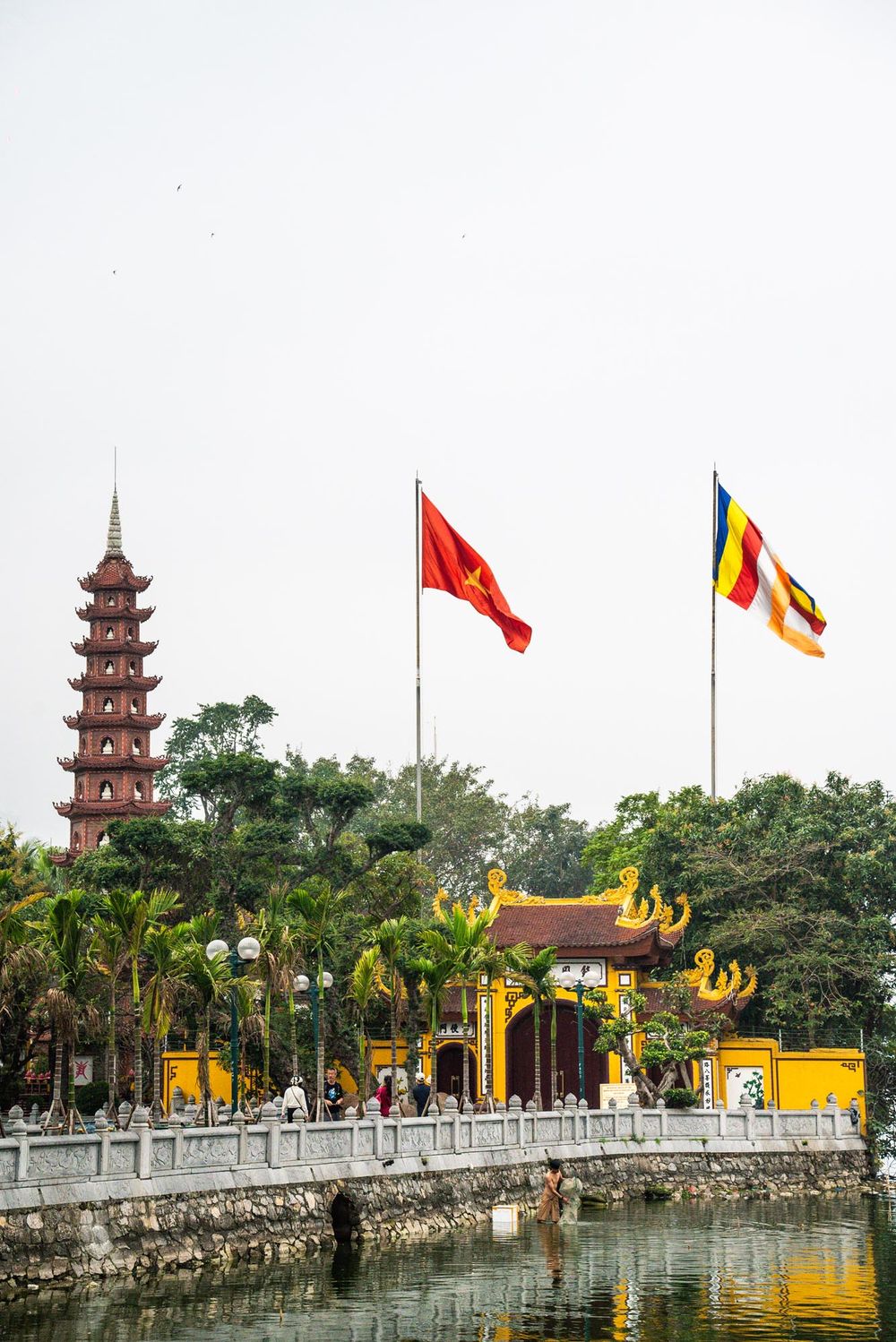 Tran Quoc Pagoda temple with Vietnamese and Buddhist flags flying beside a lake in Hanoi, featuring a tall tower and yellow buildings.