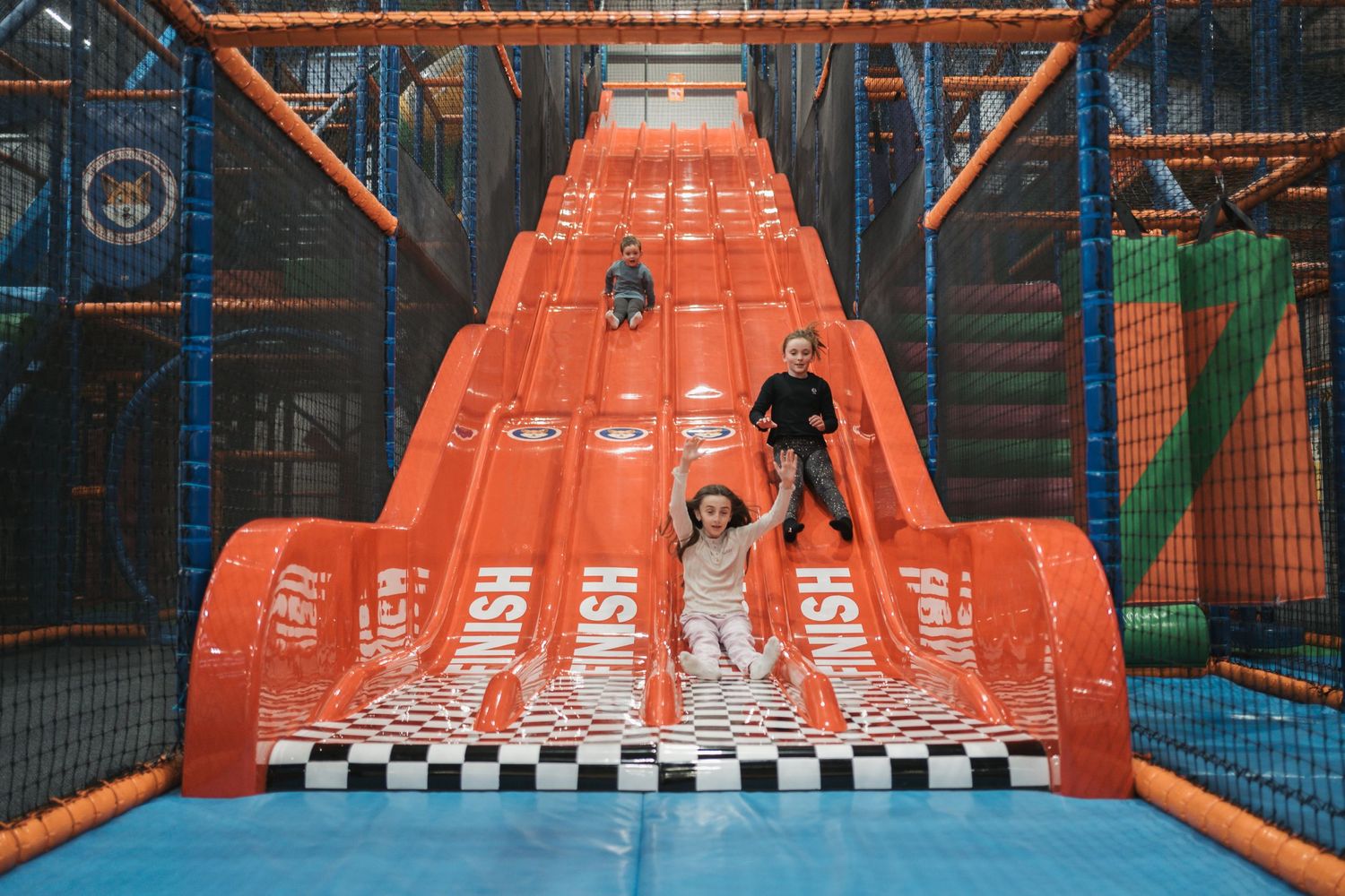 Steep orange slide with checkered landing pad inside indoor playground area with blue mat and safety netting.