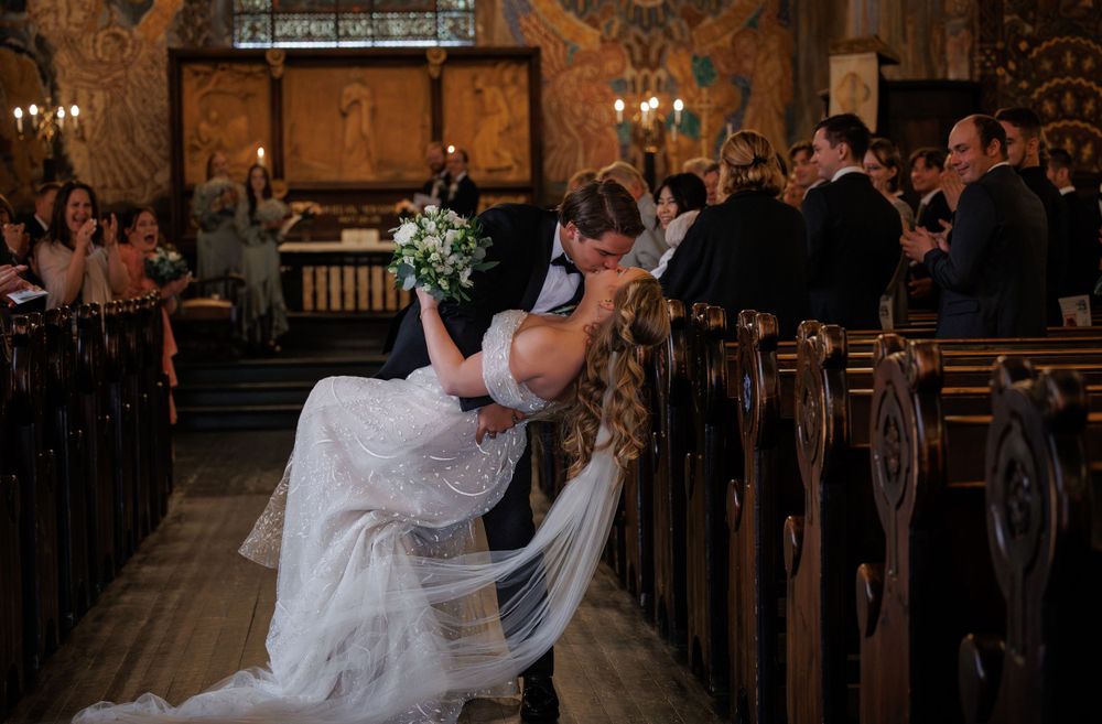 A couple shares a romantic kiss in a wooden pew during their wedding ceremony in a historic church with warm lighting.