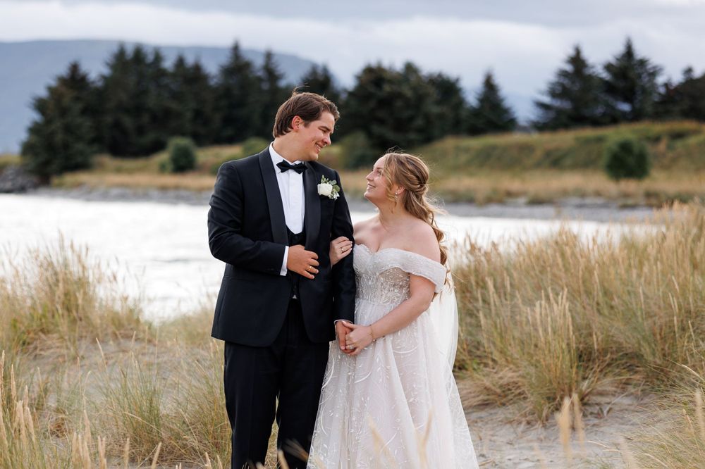 A couple in formal wedding attire walk together on a sandy beach path surrounded by tall grasses and evergreen trees.