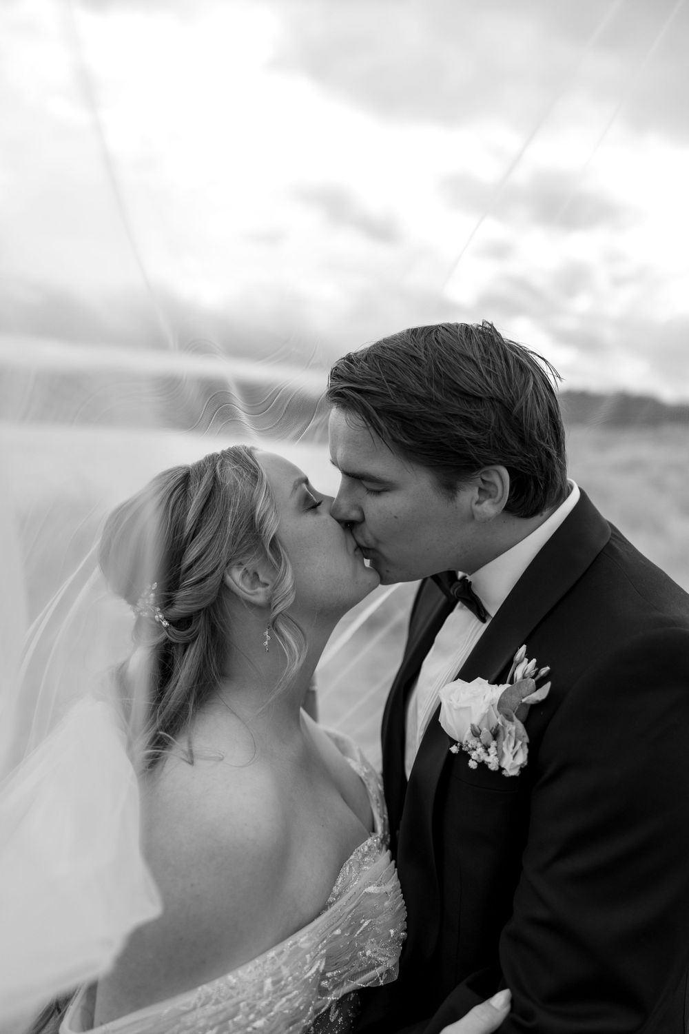 Romantic black and white wedding photo of a couple sharing a kiss under a flowing veil and cloudy sky.