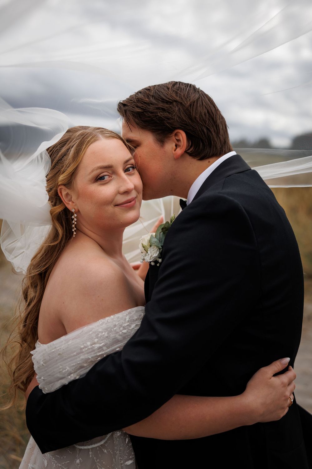 A romantic wedding couple embraces outdoors under cloudy skies with the bride's veil flowing in the wind.