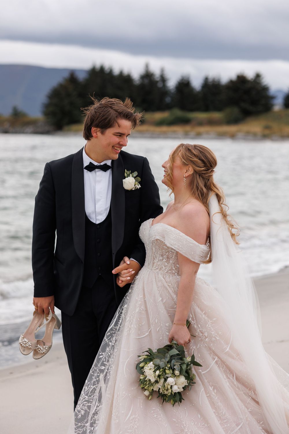 A couple in wedding attire shares a joyful moment walking along a scenic beach with mountains and evergreens in the background.