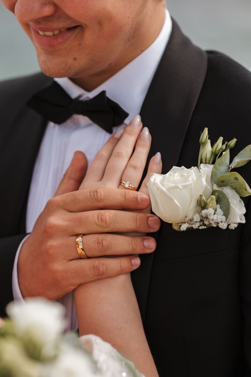 Close-up of a formal wedding attire showing a boutonniere with white rose, black tuxedo, and groom's wedding ring.