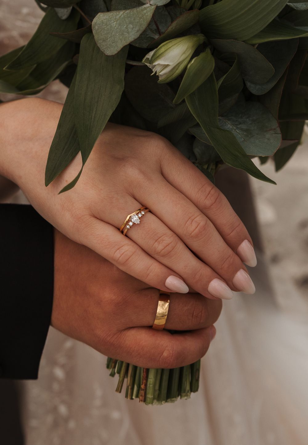 Close-up of wedding rings and white manicured hands holding a flower bouquet with green leaves.