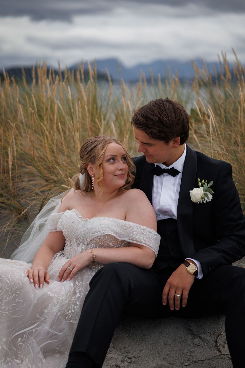 A romantic wedding couple sits together in beach grass with moody clouds in the background.