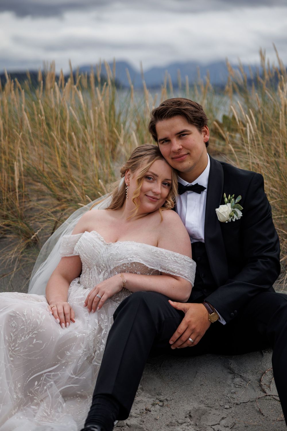A newly married couple sits together among tall beach grass on a cloudy day.