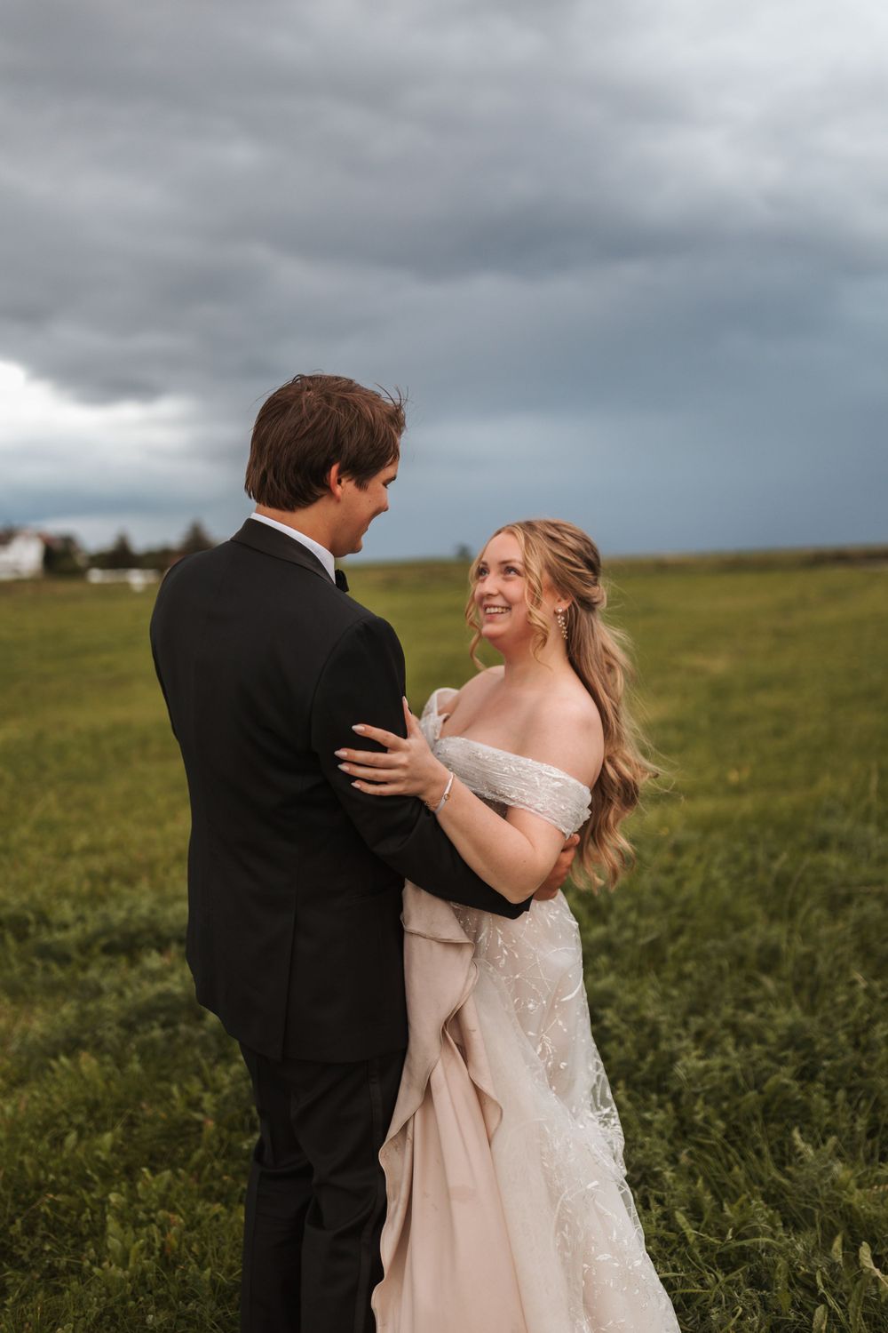 A couple in wedding attire embraces in a field under dramatic stormy skies.