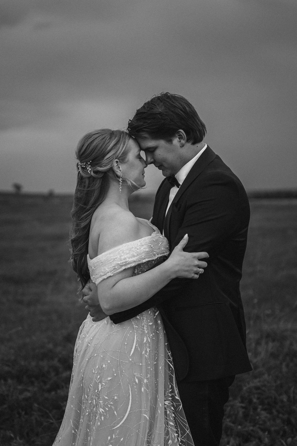 A romantic black and white outdoor wedding photo captures a tender moment between newlyweds in formal attire.