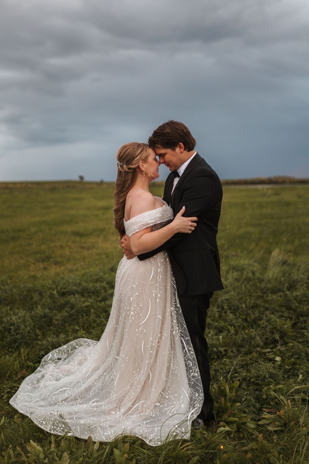 A wedding couple embraces in a romantic moment on grassy field under stormy skies.