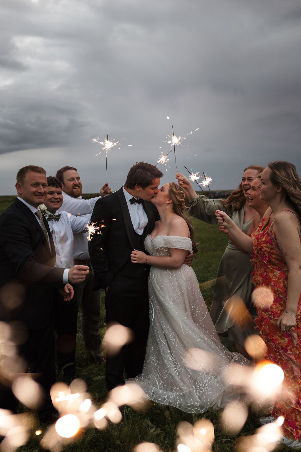 Wedding celebration in a field at dusk with sparklers creating a magical atmosphere under stormy skies.