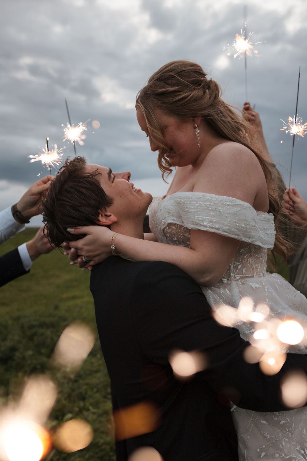 Newlywed couple embraces outdoors at sunset with sparklers creating magical bokeh light effects around them.
