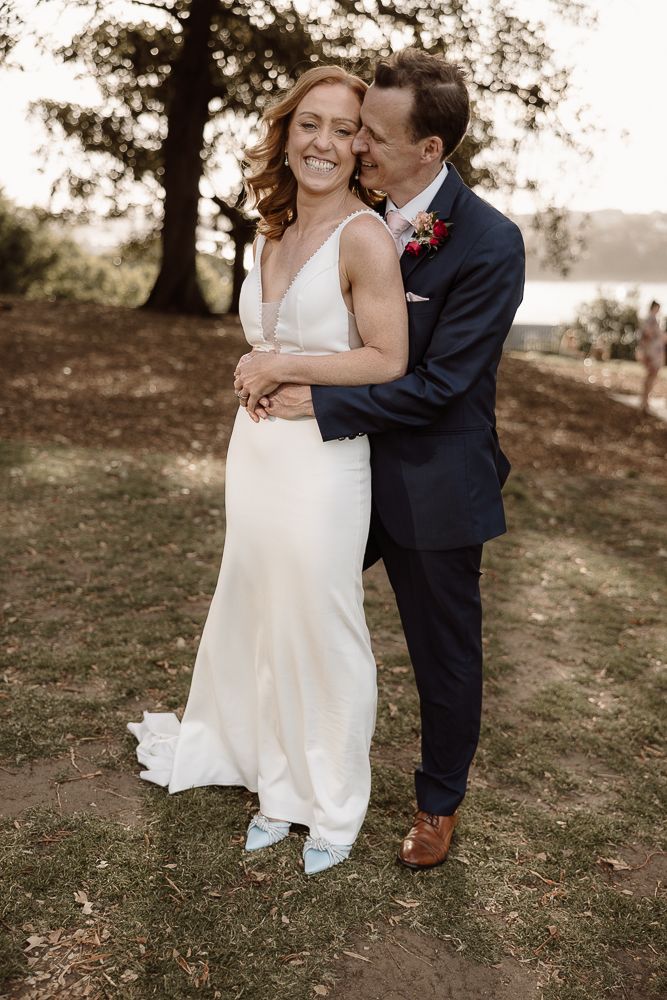 A newlywed couple embrace outdoors in a natural setting, the bride in white dress and the groom in navy suit.