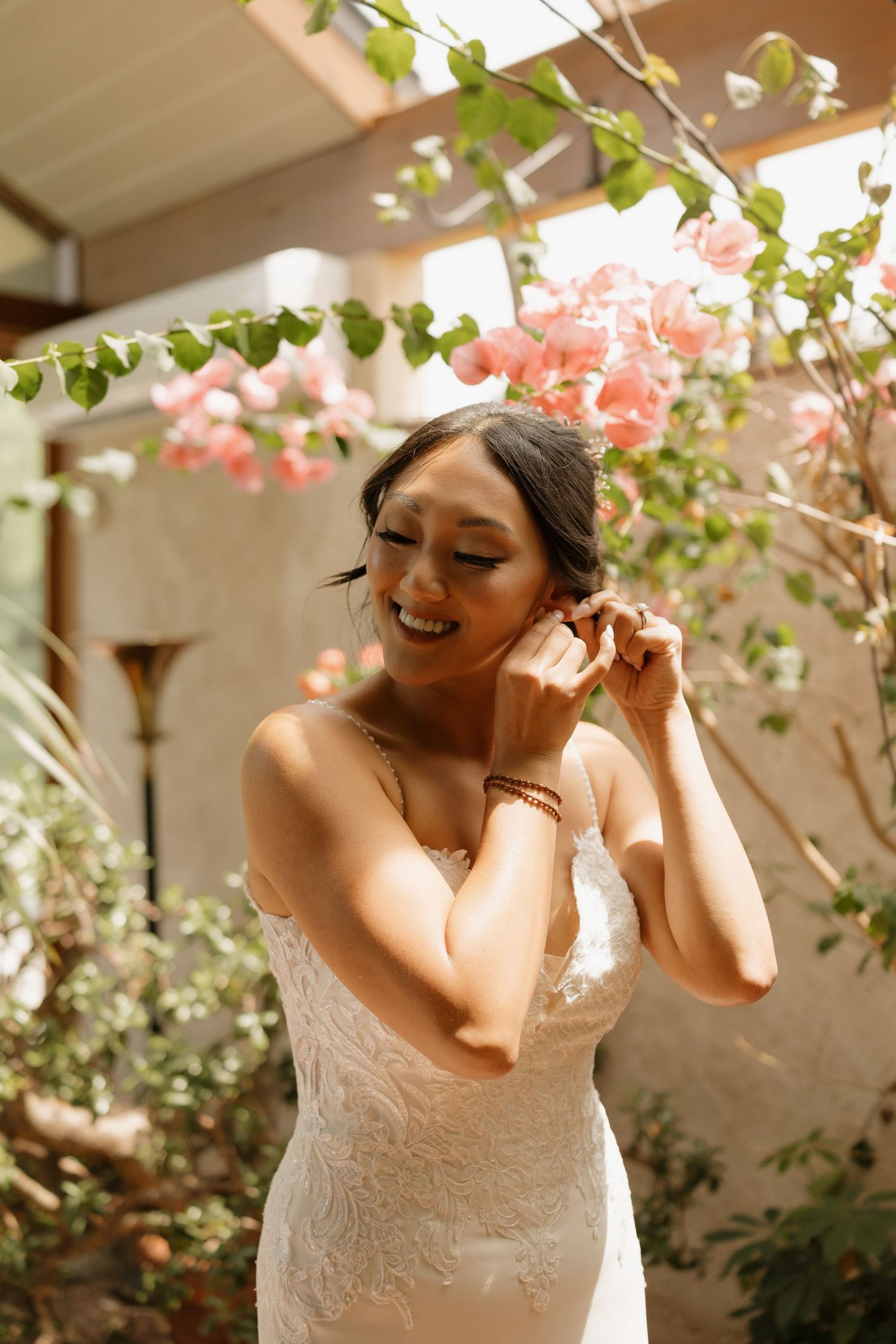 Bride wearing a white lace dress laughs joyfully during an outdoor photo session with pink flowers in the background.