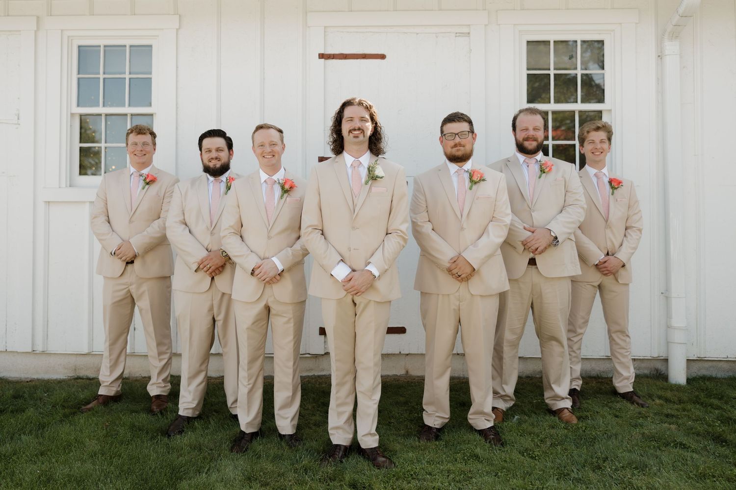 Wedding party in matching beige suits stands against white building exterior for formal group photo.