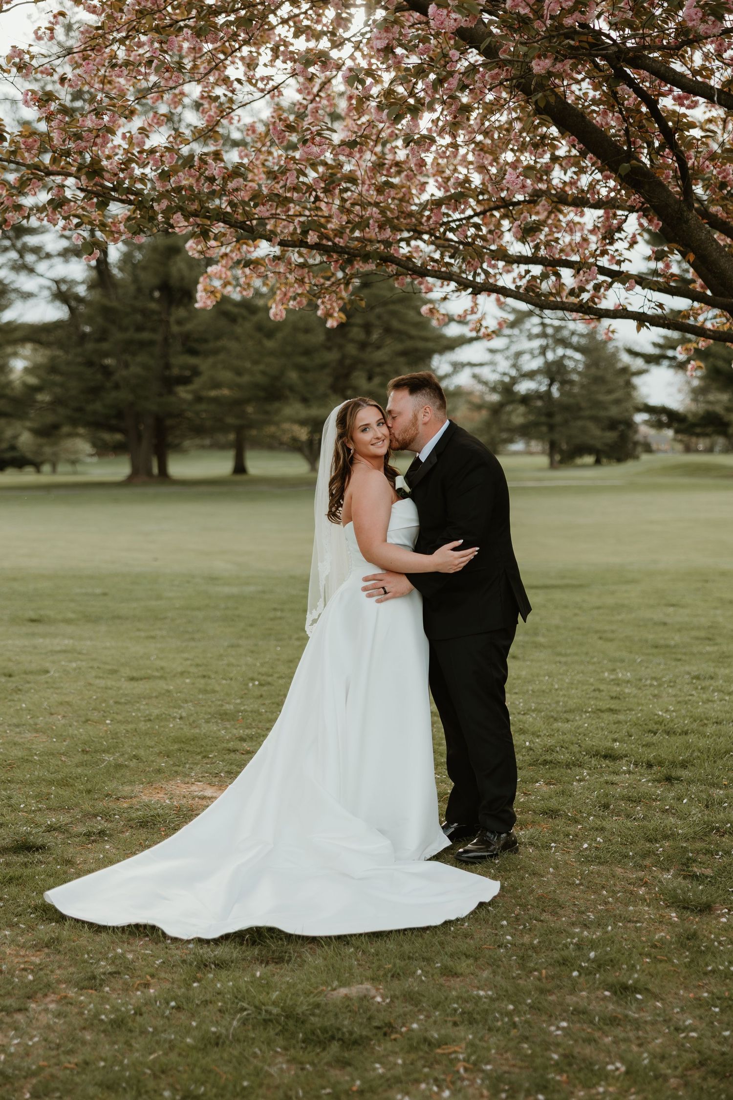 A couple shares a romantic moment under blooming cherry blossoms on a lush green lawn during their wedding day celebration.