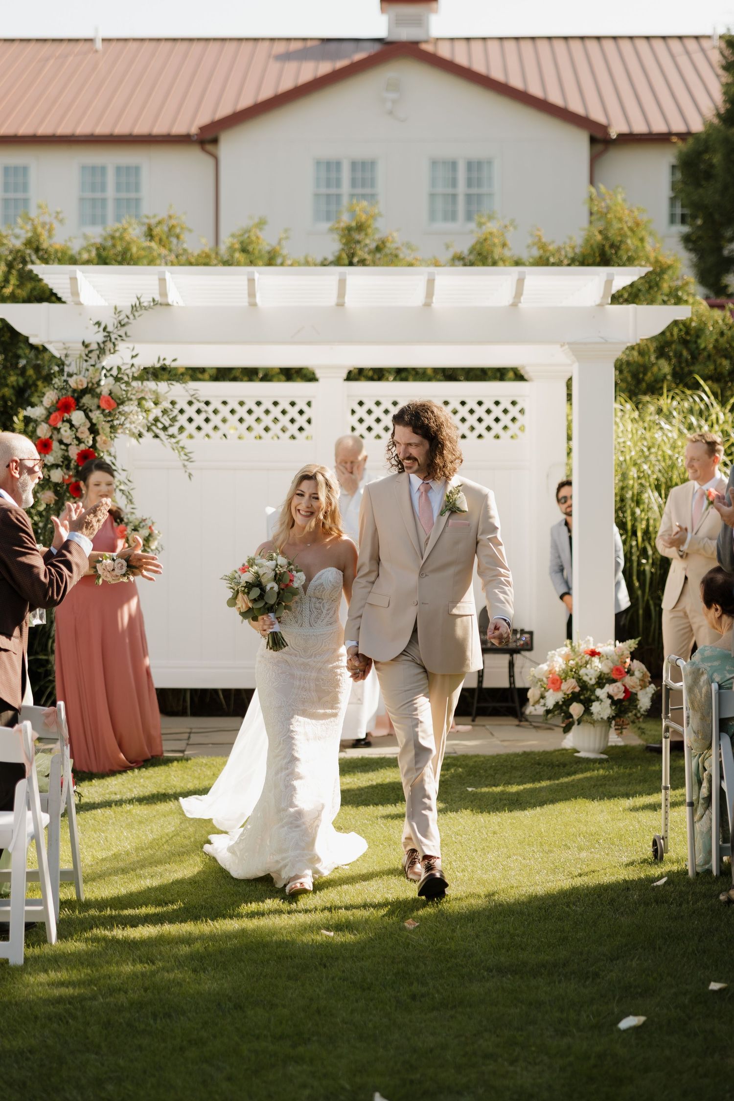 A wedding couple walks down an outdoor aisle under a white pergola surrounded by flowers and coral-dressed attendants.