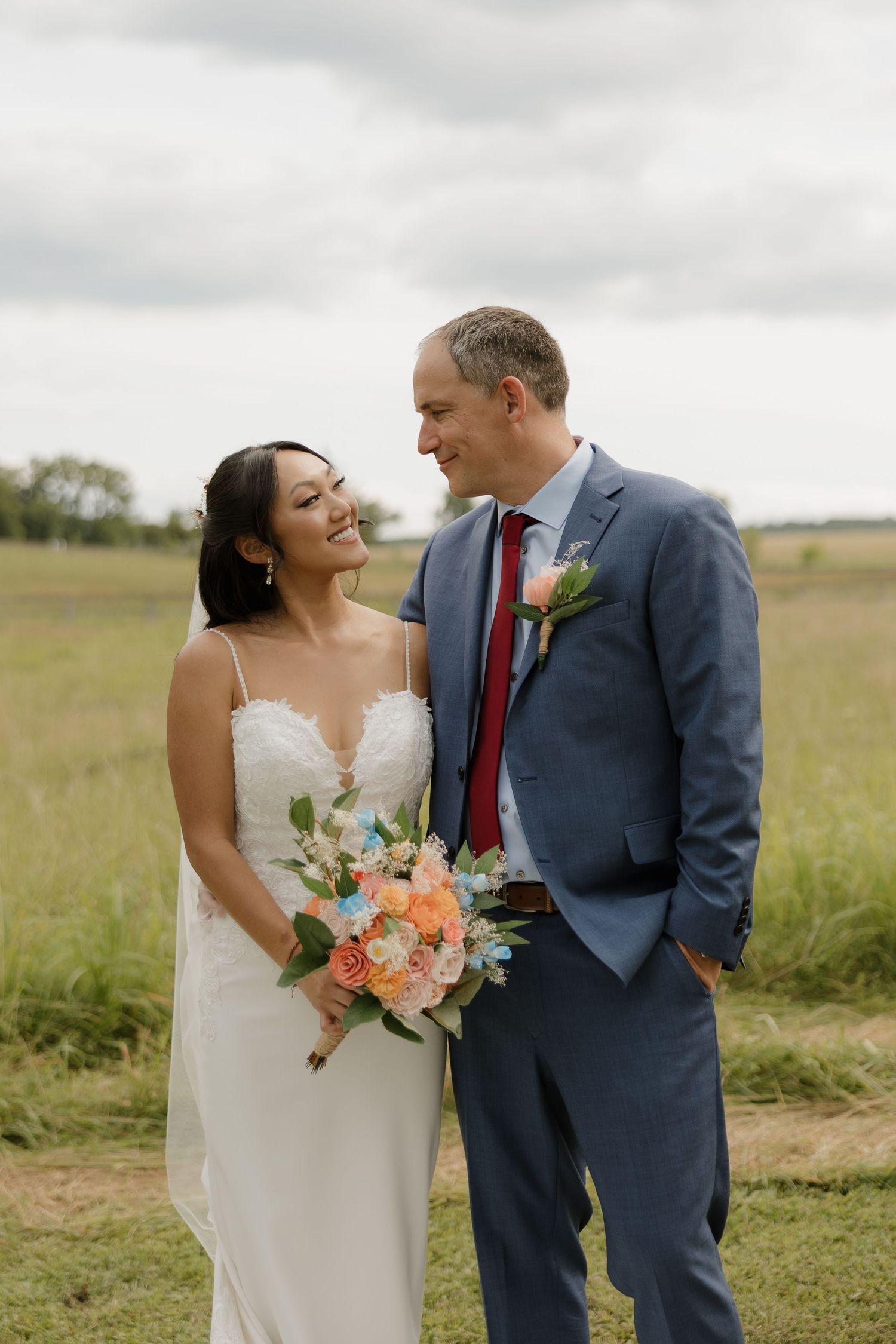 A romantic wedding couple stands in a grassy meadow, with the bride holding a peach and coral bouquet.