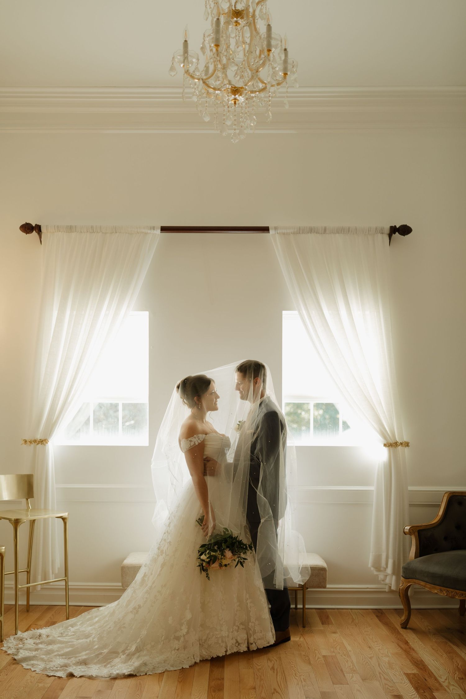 A dreamy wedding couple dances together in a bright white room with soft natural lighting and flowing white curtains.