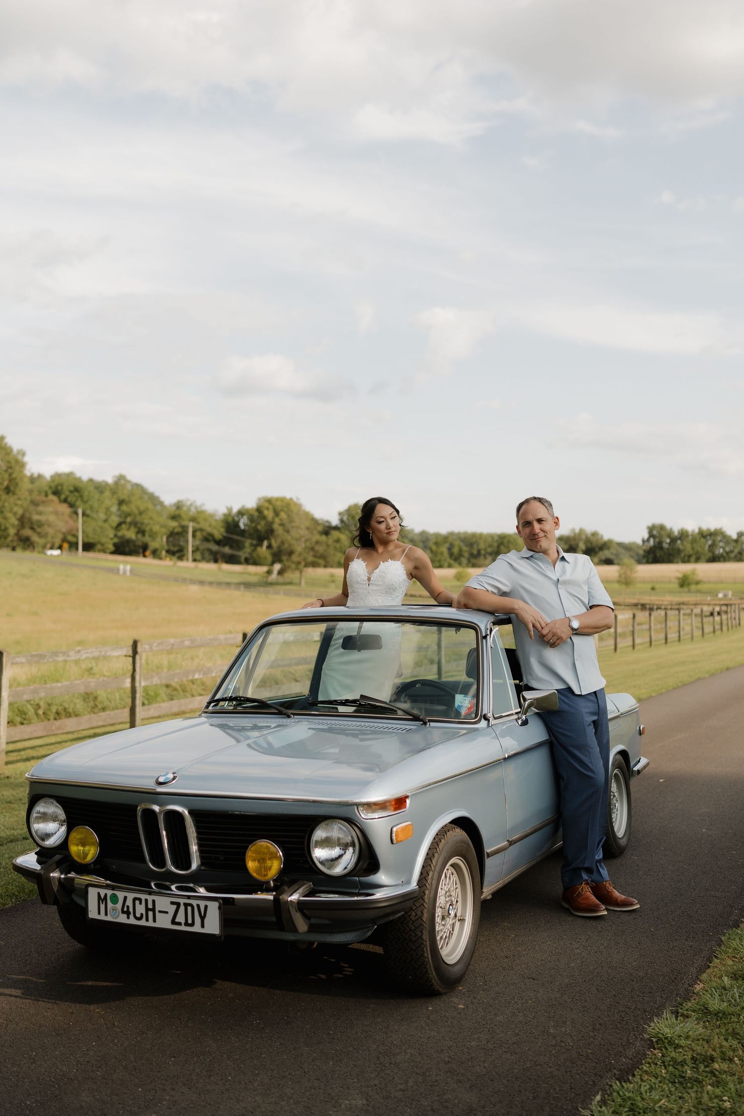 Couple posing with a vintage silver BMW convertible on a rural country road during a sunny afternoon.