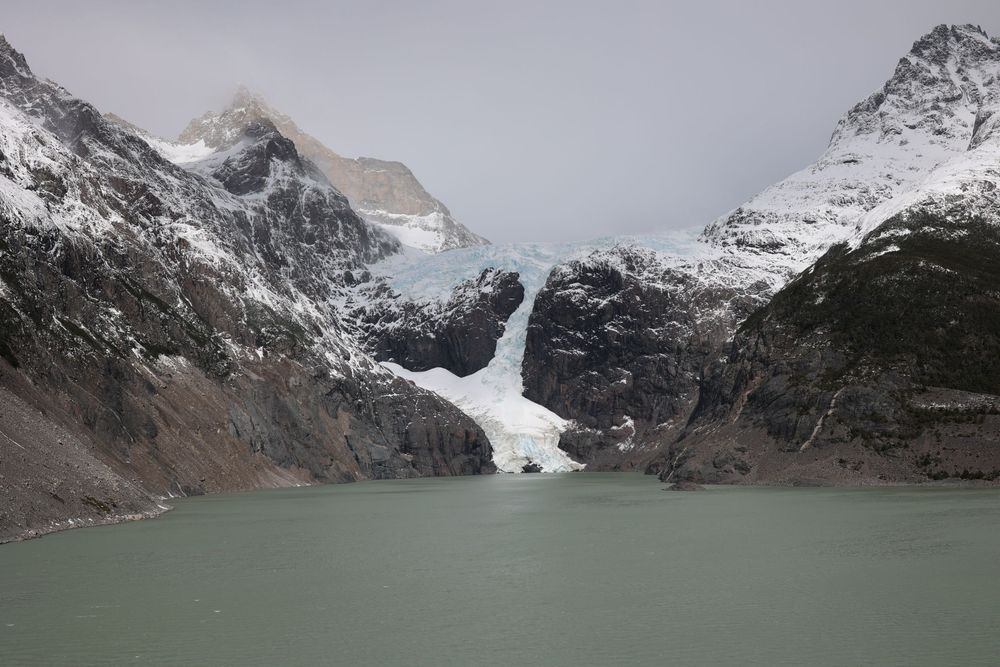 Majestuoso glaciar helado que desemboca en un tranquilo lago rodeado de escarpadas montañas nevadas bajo un espectacular cielo nublado.