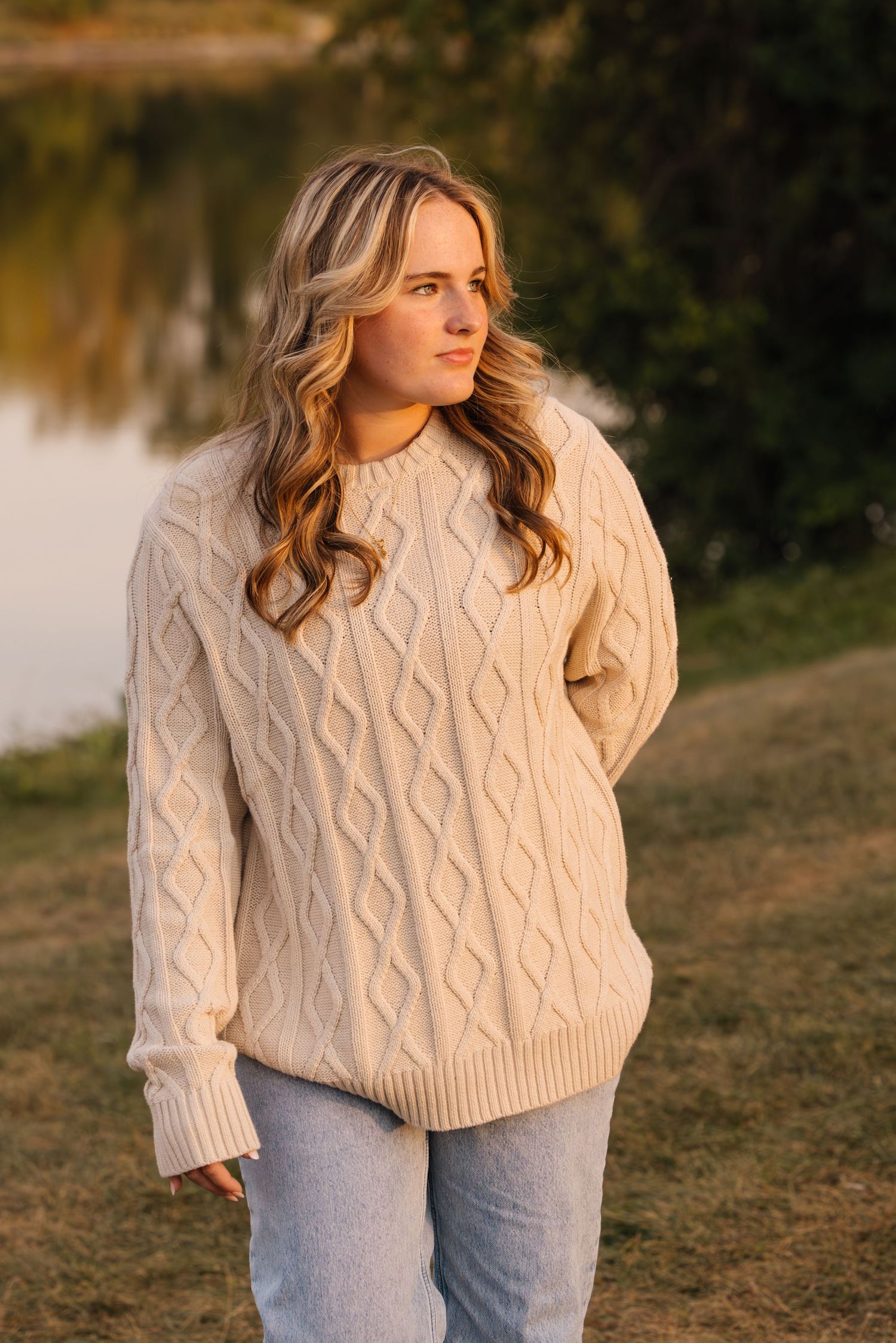 A person wearing a cream cable knit sweater and light jeans stands near a lake during golden hour.