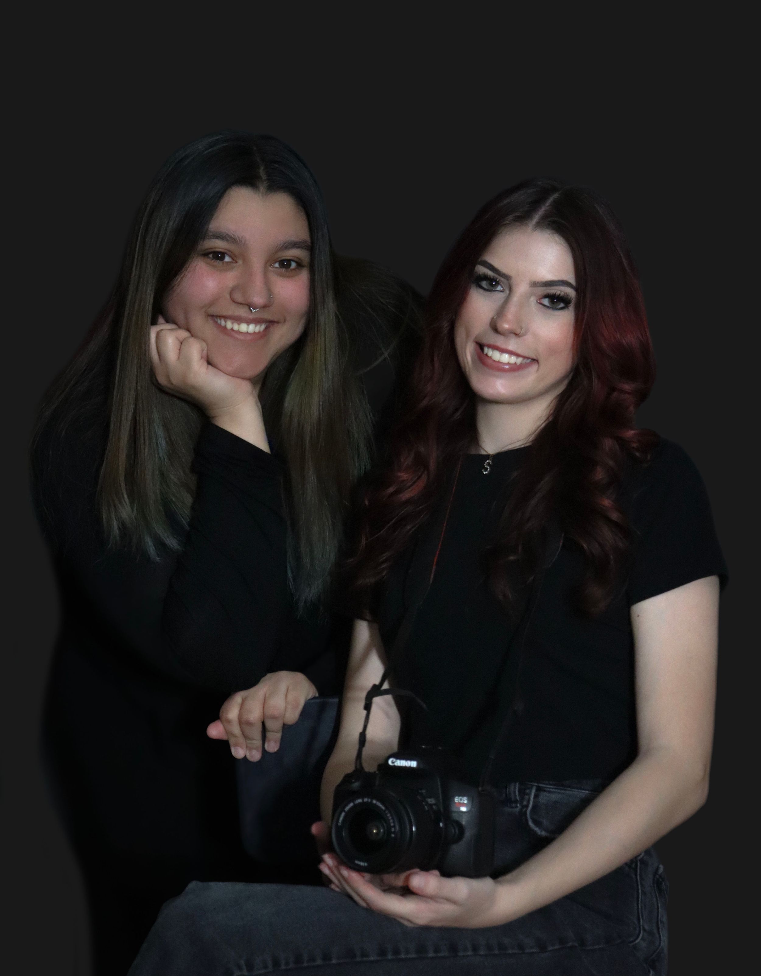 Portrait of two friends in black outfits smiling at camera with one holding a professional DSLR camera.