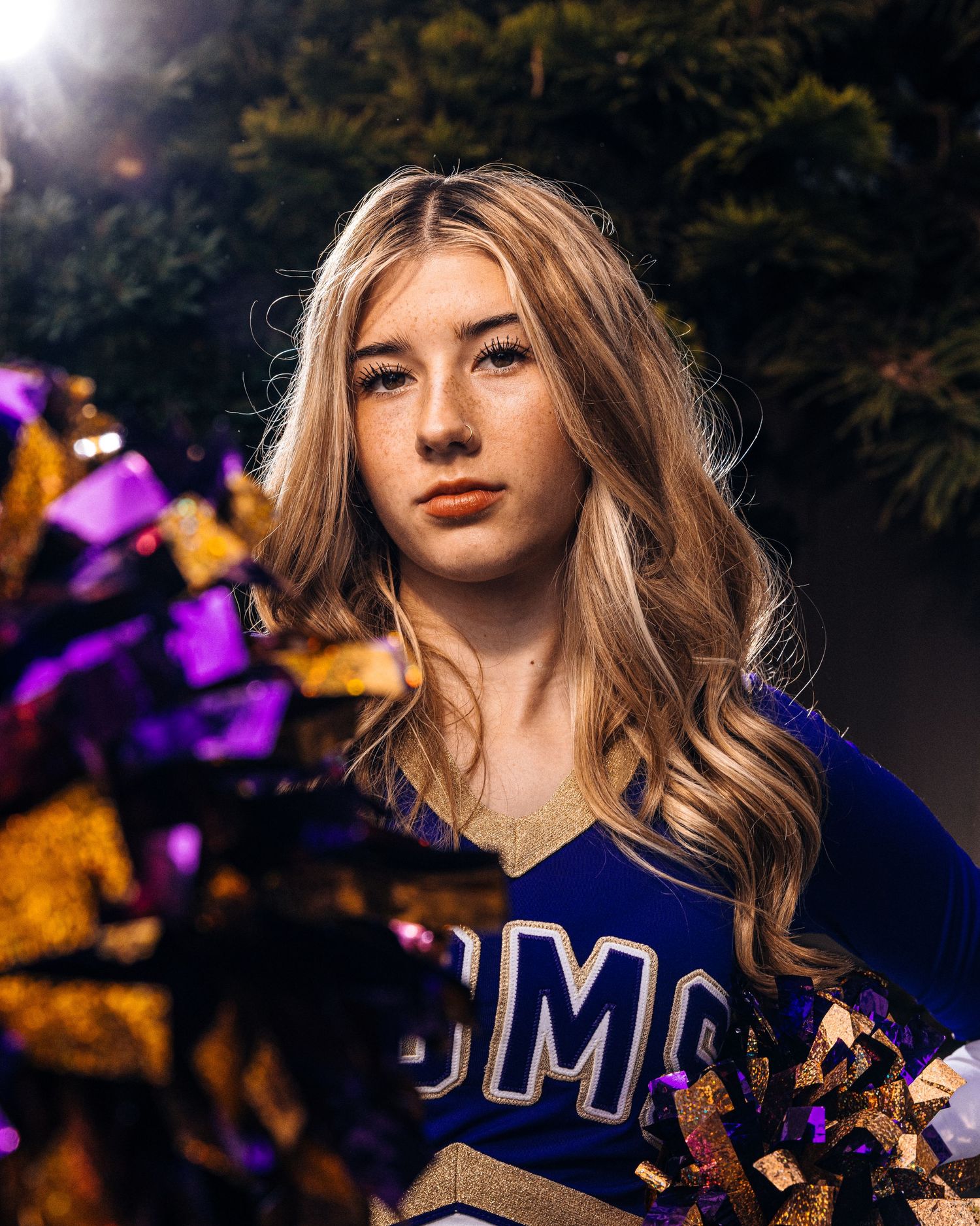 A cheerleader wearing a blue uniform poses outdoors with autumn foliage in the background.