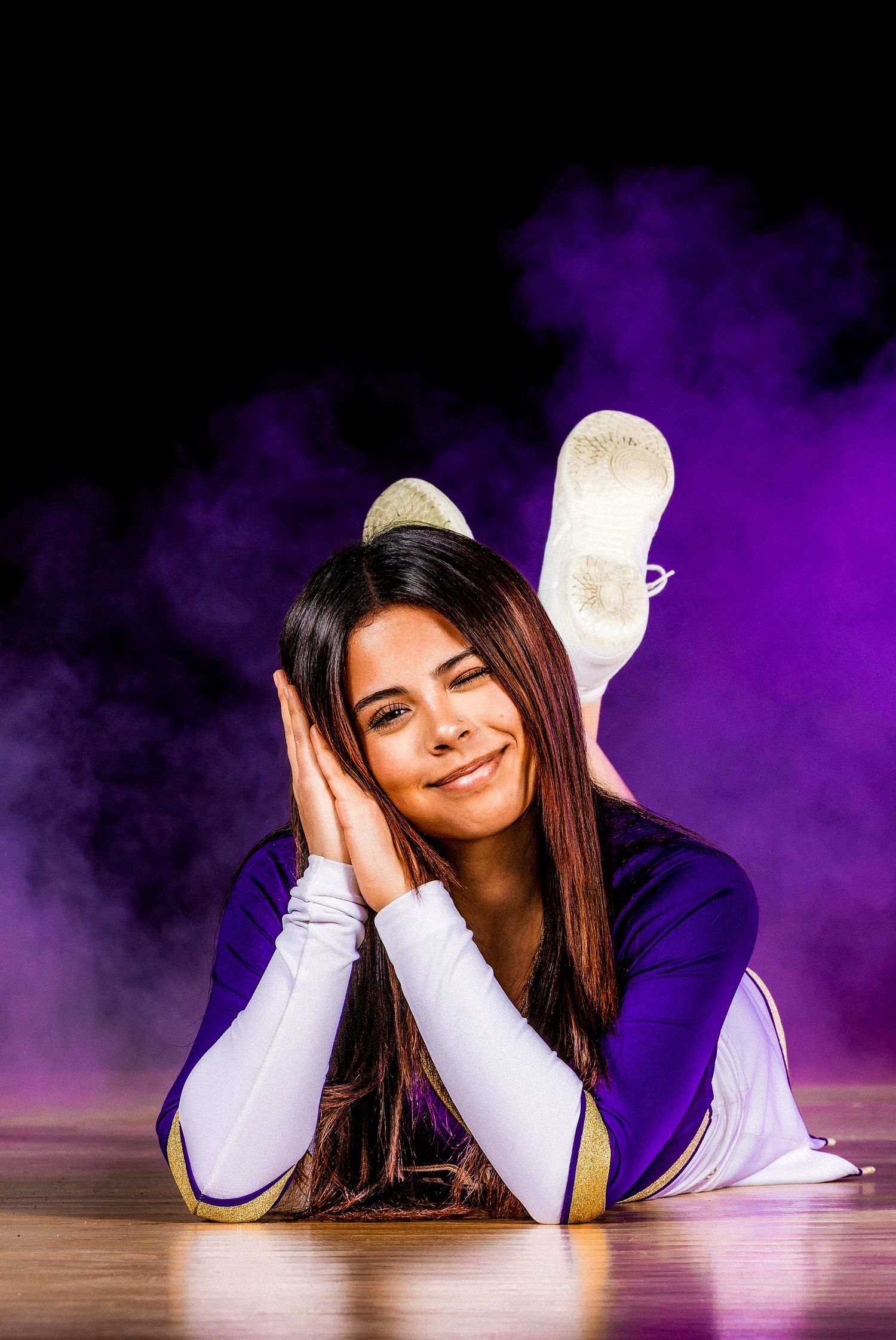 A cheerleader in purple uniform lying on wooden floor with surrounding purple smoke effects.