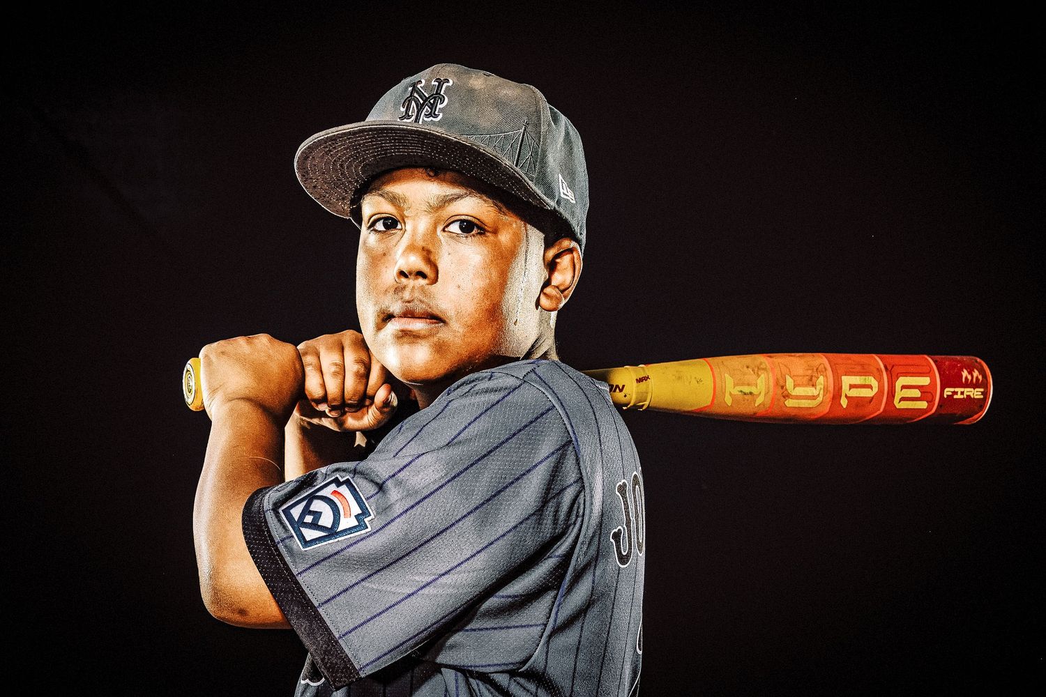 Baseball player in New York Yankees uniform poses with wooden bat against dark background.