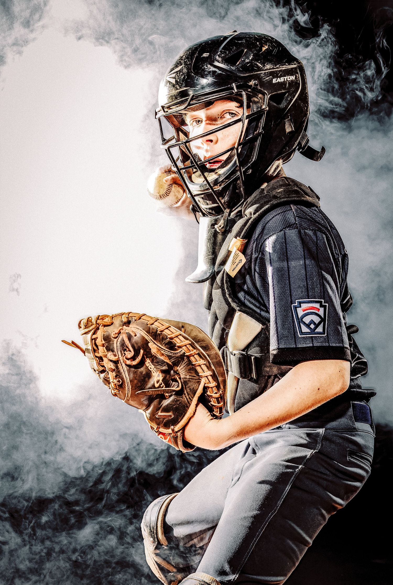Baseball catcher in black protective gear and brown mitt poses against dramatic smoky background.