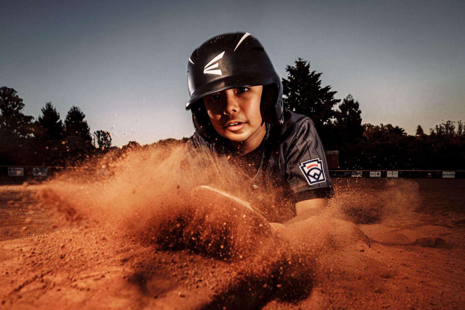 Motocross rider kicks up orange dirt while speeding through a dusty track at sunset.
