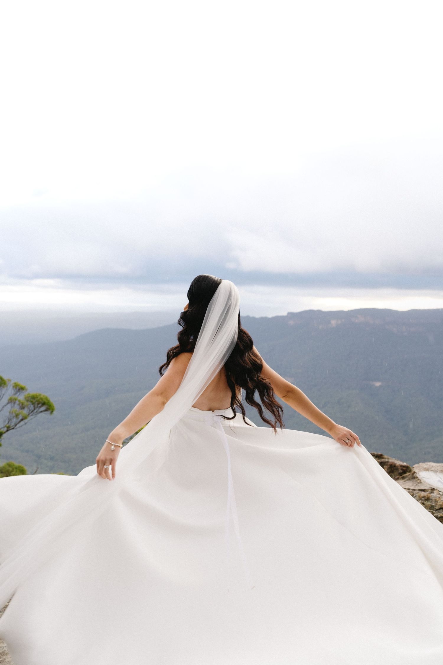 A bride in a flowing white wedding dress spins atop a mountain overlook with cloudy mountains in the background.