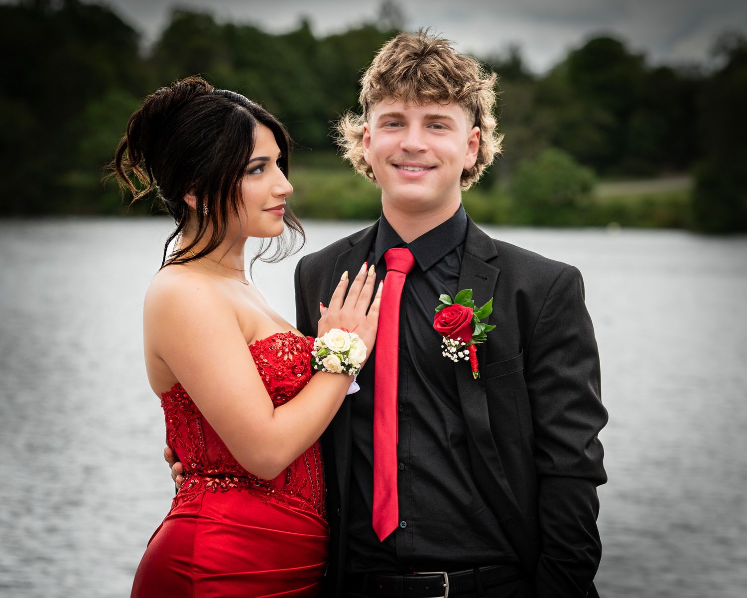 Romantic formal couple poses by lake with woman in red dress and man in black suit with red tie.