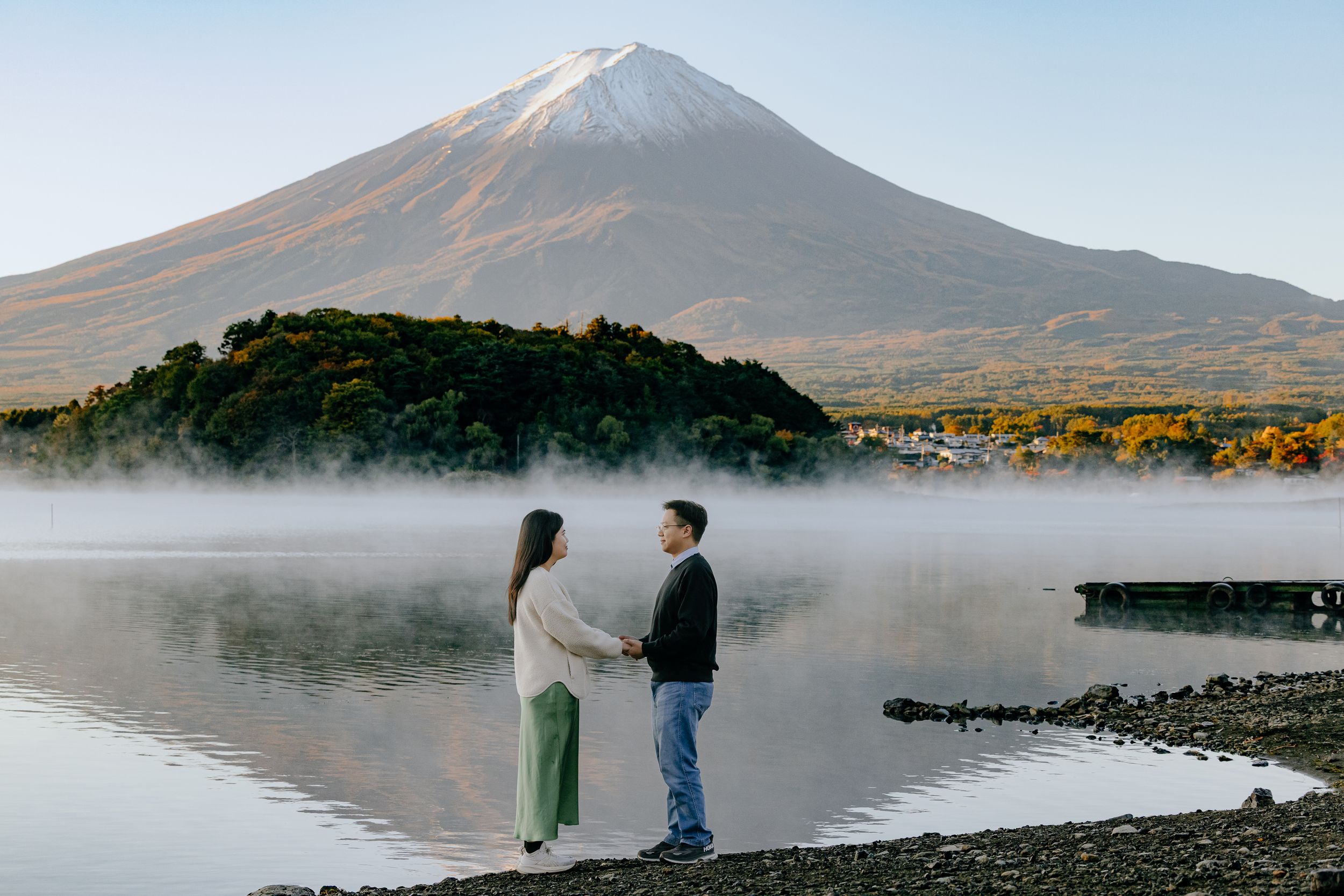 Two people stand on the misty shore of a mountain lake during sunrise with a snow-capped peak in the distance.