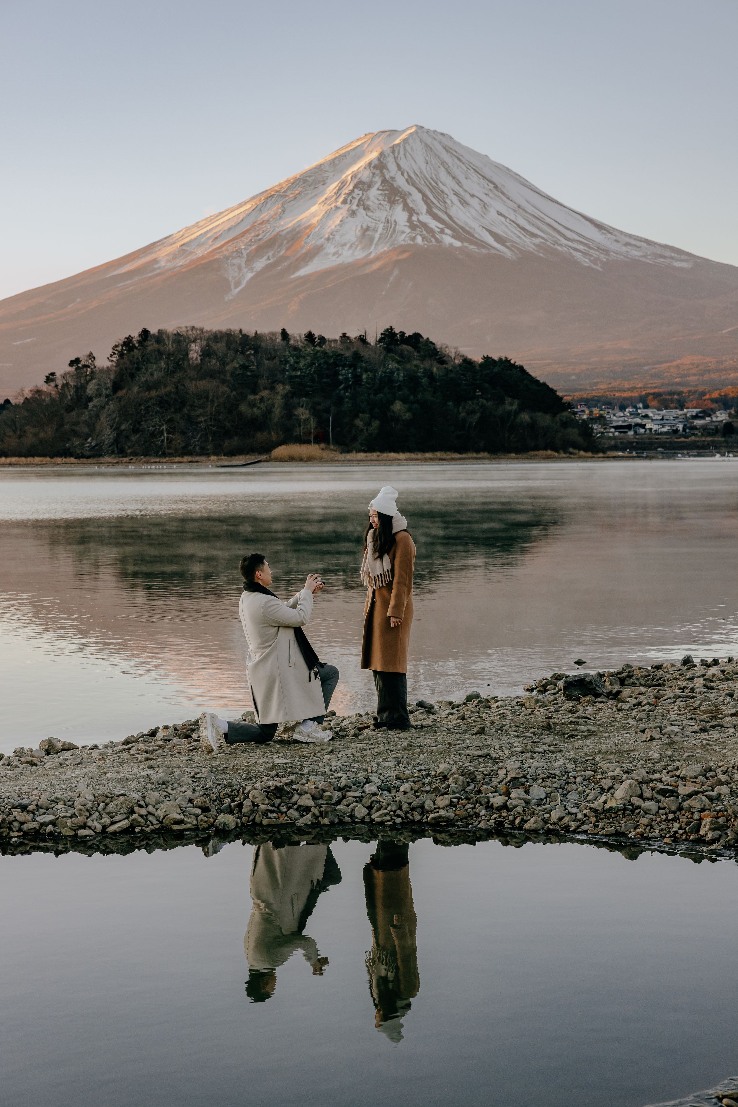 Artist painting at tranquil lakeside with snow-capped Mount Fuji reflecting in calm waters at sunset.