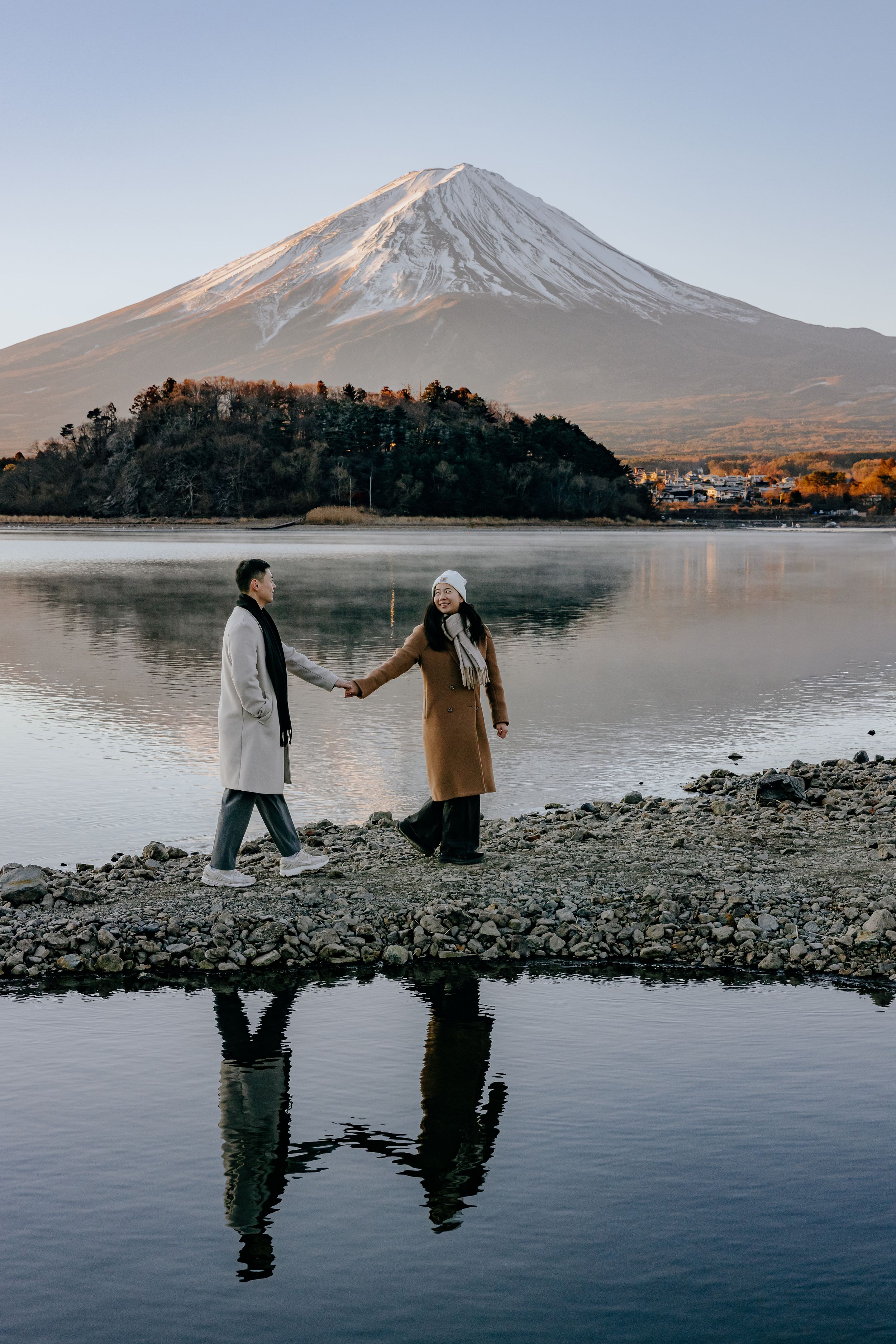 Two people walking along a serene lakeshore with Mount Fuji's snow-capped peak rising majestically in the background.