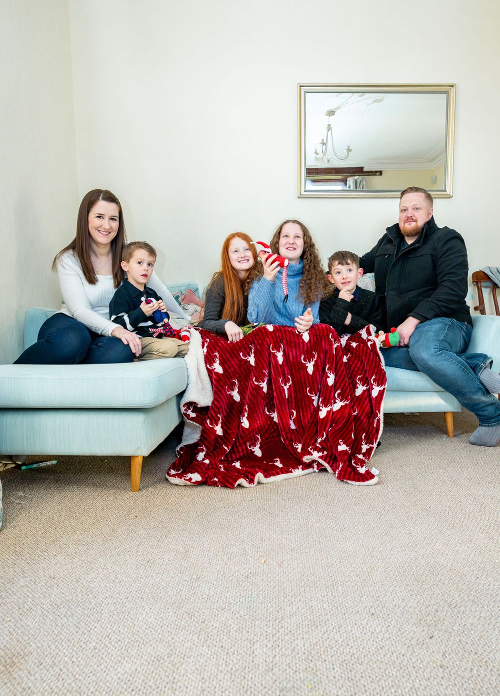 Family sitting together on gray sectional sofa covered with red reindeer patterned blanket in living room.