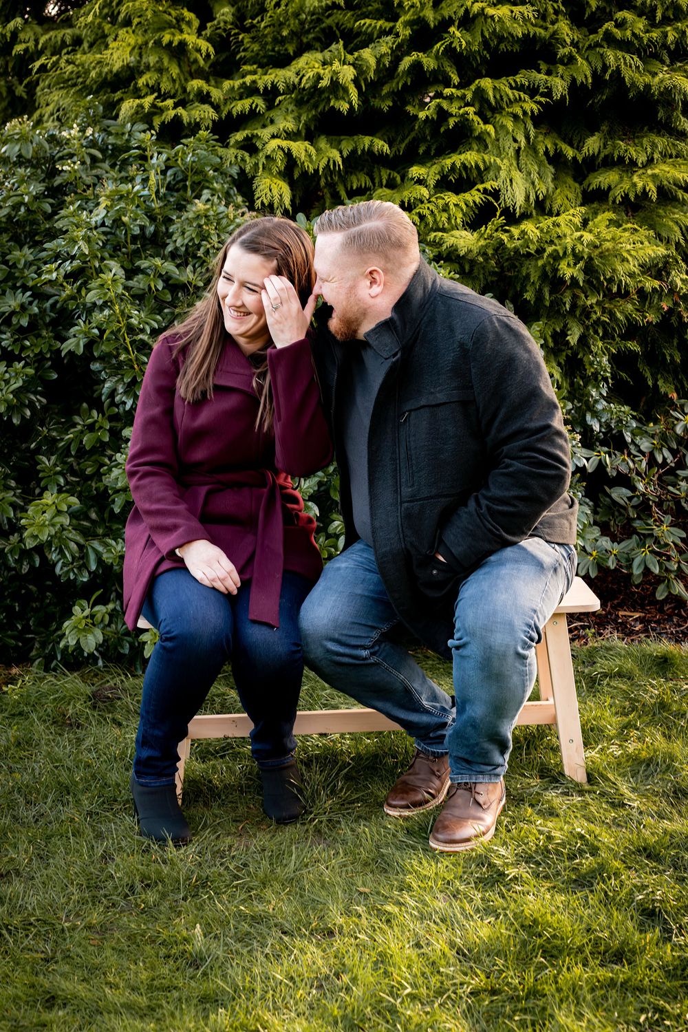 Couple sharing an intimate moment while sitting on wooden bench against evergreen hedge in backyard garden setting.