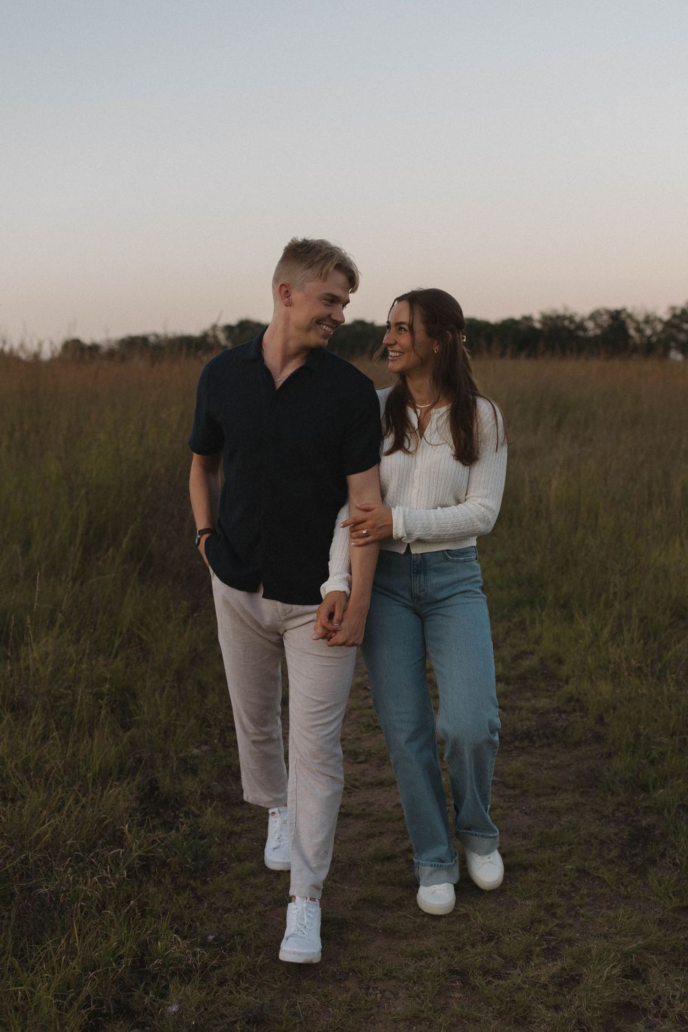 Couple laughs together while walking through a rustic field during sunset.