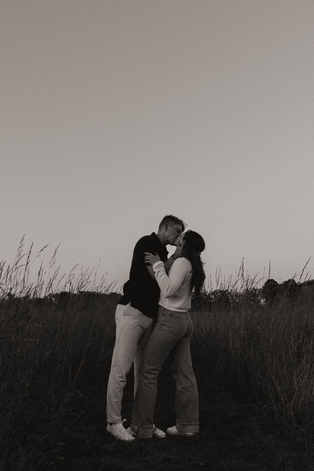 Silhouetted couple embraces in a field during twilight with tall grass in the background.
