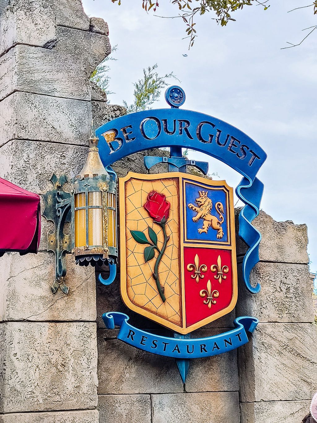 Decorative restaurant sign featuring a gold and blue heraldic shield with fleur-de-lis emblems on a stone castle wall.