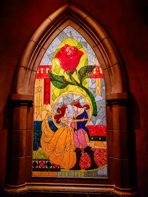 Stained glass window depicting Beauty and the Beast dancing with a red rose in an ornate Gothic church-style frame.