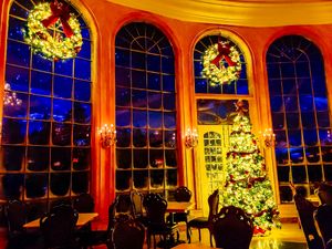Elegant dining room with illuminated Christmas tree and wreaths glowing against arched windows in warm golden light.