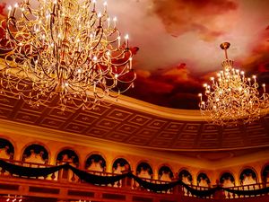 Ornate crystal chandeliers hang from a ceiling with dramatic red clouds painted across its baroque architectural details.