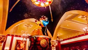Colorful acrobatic circus performer balances on a beam under ornate red and gold ceiling decor and chandelier lighting.