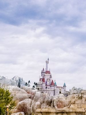 Beauty and the Beast castle with pink and blue spires rises above rocky terrain against a cloudy sky at Disney theme park.