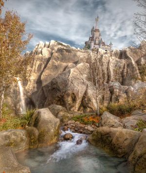 A castle perched atop rocky cliffs with a waterfall cascading down into a flowing stream against a dramatic cloudy sky.